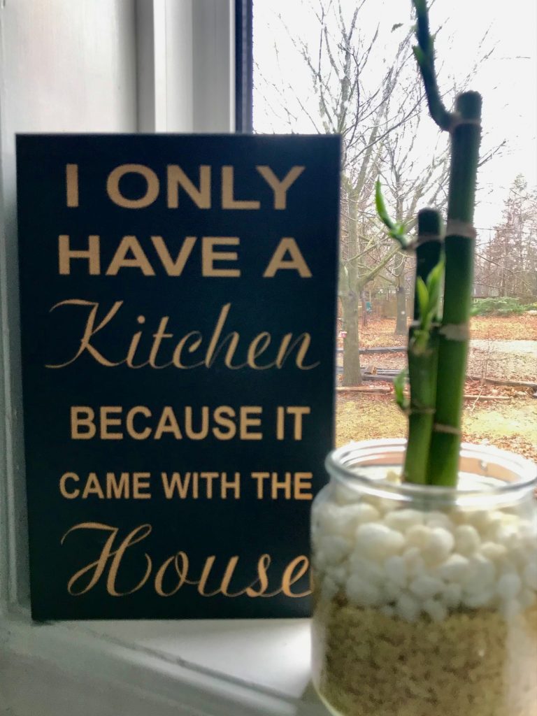 Small wood sign that says I only have a kitchen because it came with the house beside a small lucky bamboo plant.